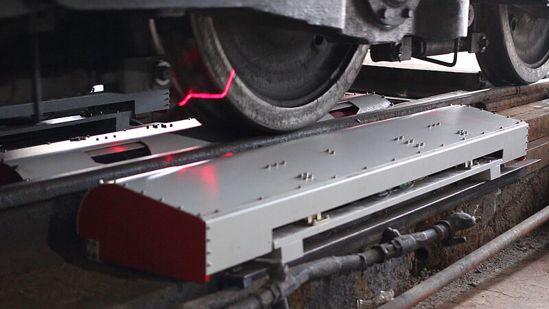 A photo showing a metallic box embedded in the track. Above it, the wheels of a train can be seen and the pink laser beam coming out of the box.