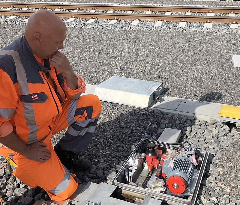 Maintenance technician in orange work clothes sitting in the track in front of an open case with a turnout mechanism.